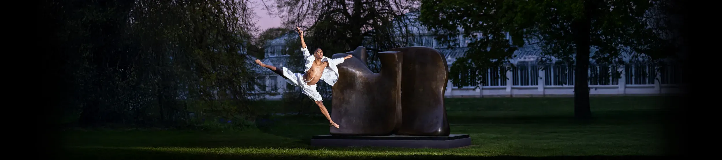 Image of a dancer leaping in front of a Henry Moore sculpture, at dusk. The Temperate House is in the background.