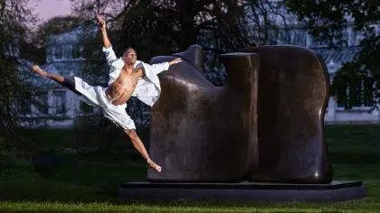 Image of a dancer leaping in front of a Henry Moore sculpture, at dusk. The Temperate House is in the background.