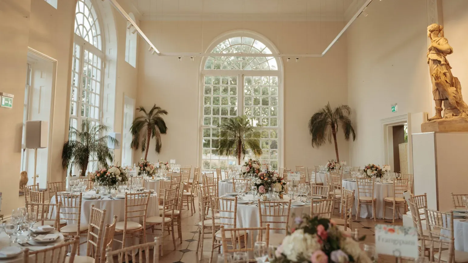 Wedding breakfast set up in a light airy hall with palm trees and ornate pink floral decorations on each table