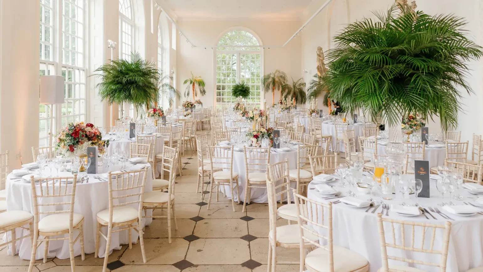 Wedding breakfast set up in a light airy hall with lush palm trees, white tablecloths and ornate rose bouquets on each table