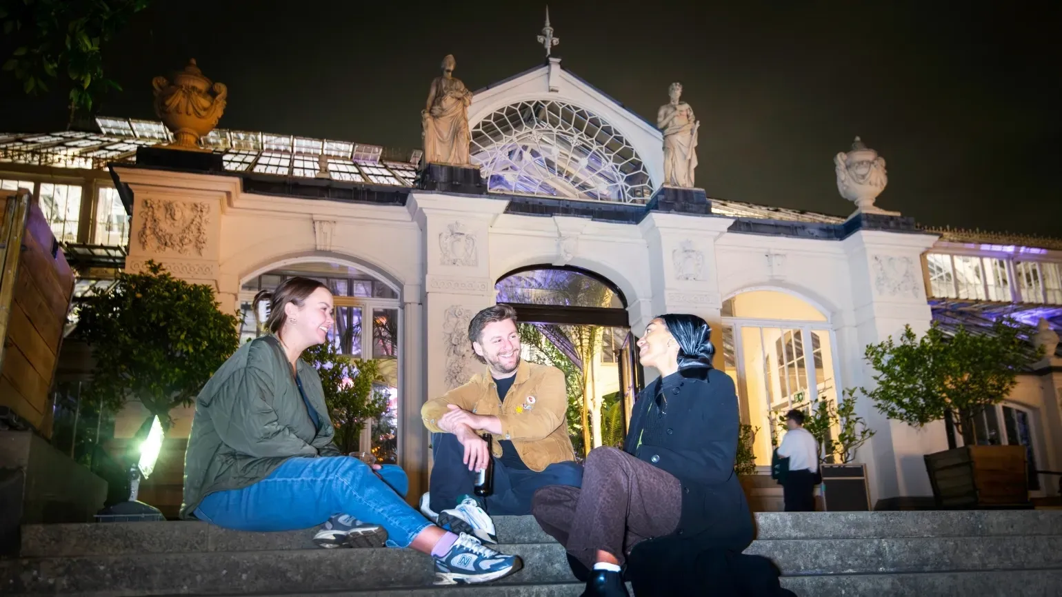 Three people sitting outside of the Temperate House at night, on the steps
