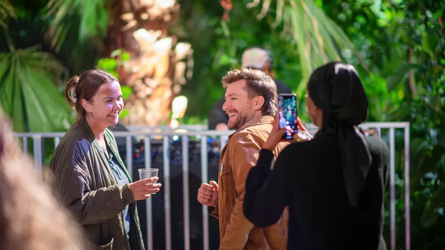 Three people dancing inside the Temperate House, with a DJ in the background. One person is capturing an image on their mobile phone.
