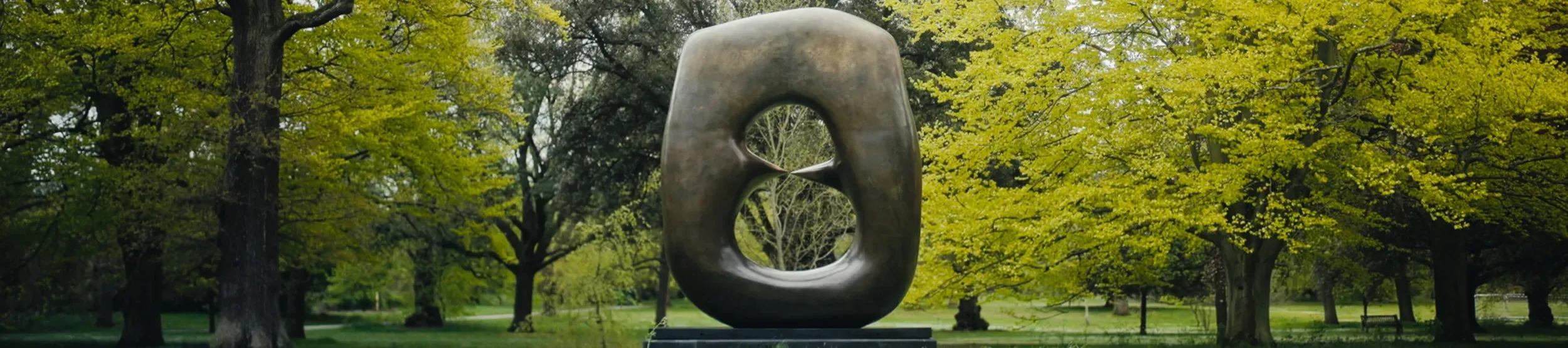 Henry Moore's monumental bronze sculpture Oval with Points in front of many green trees, with the sunlight bouncing down.