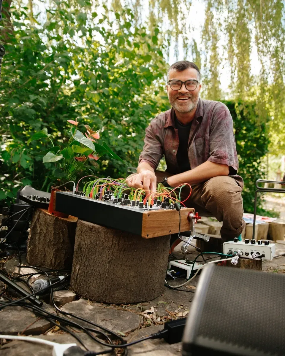 Man wearing a pair of black glasses, squatting in front of DJ equipment and smiling. There is a tree and green foliage in the background.
