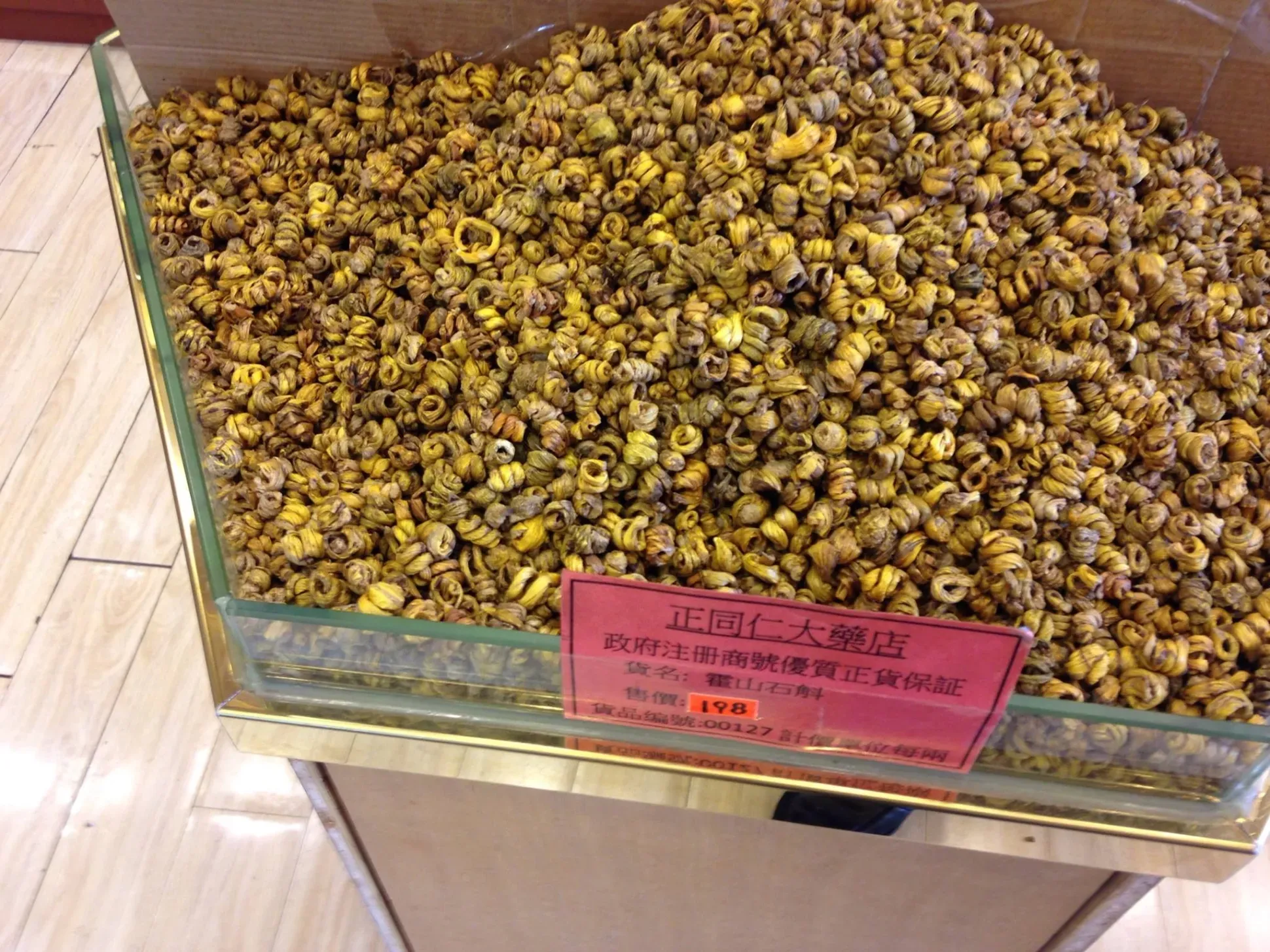 A glass display case filled with dried, curled yellow-brown Dendrobium stems for sale, with a red price label written in Chinese characters.
