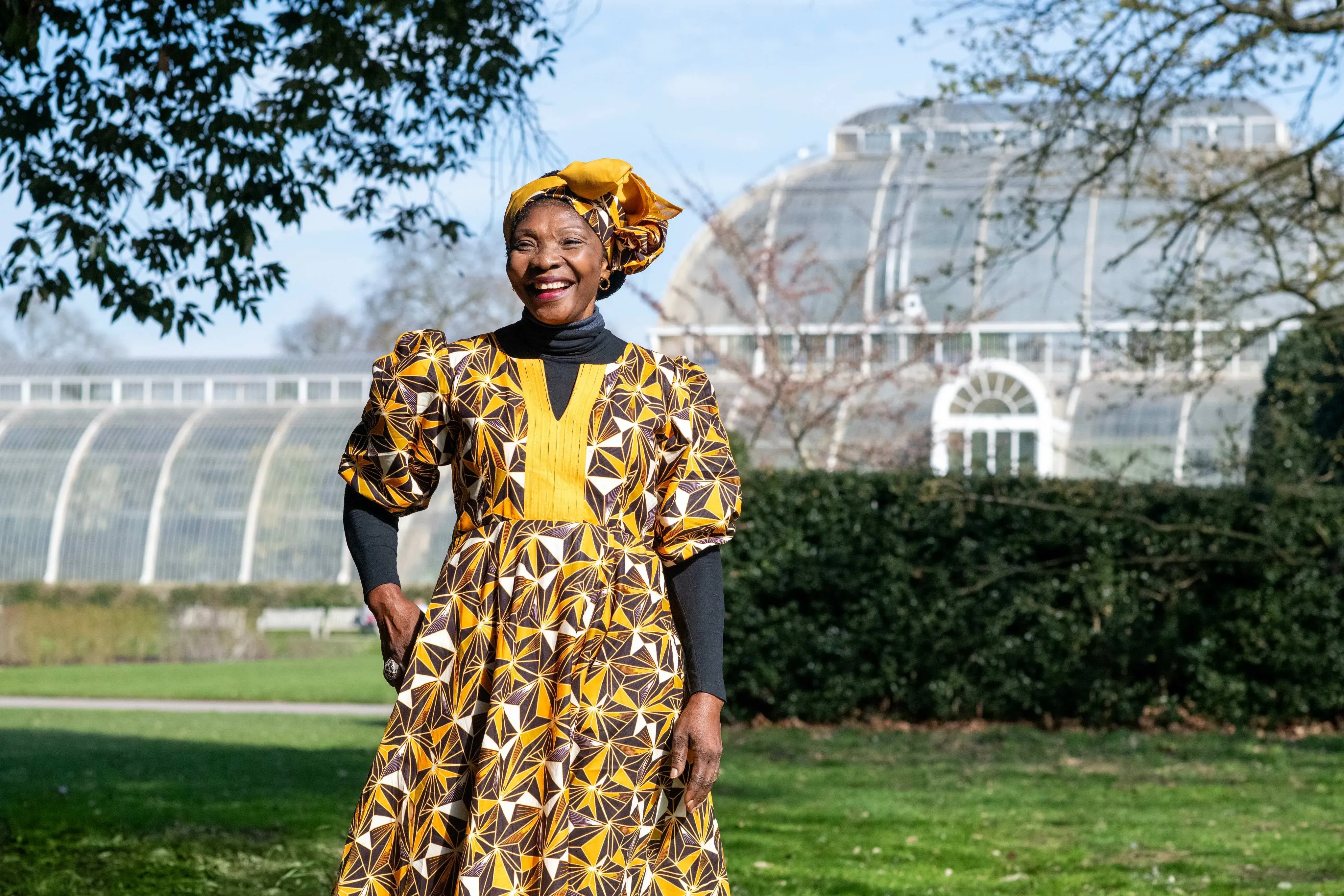 Cameroonian woman in yellow and brown patterned traditional dress and headwrap, smiling in front of a large Victorian greenhouse