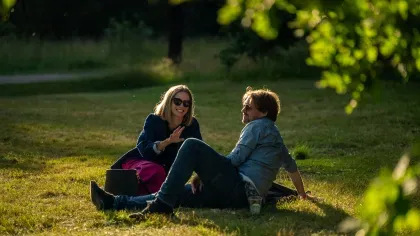 A man and woman lie on a lawn in evening light, smiling and talking