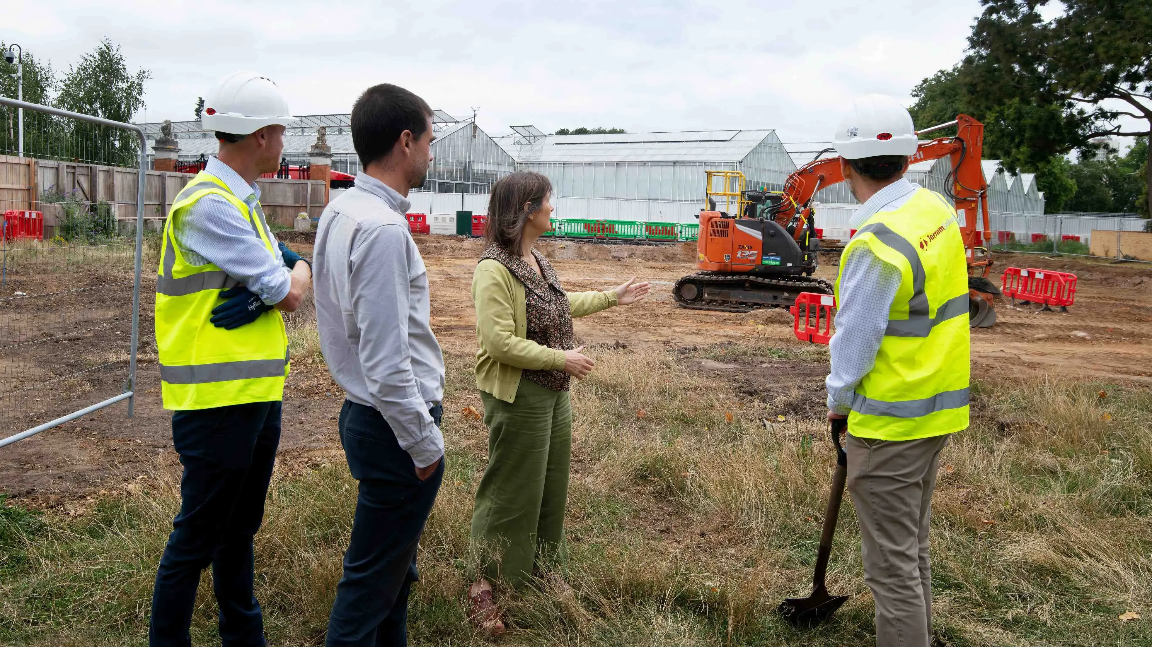 Four people stand in a construction site, two in hi vis vests, one holding a shovel, one woman gesturing