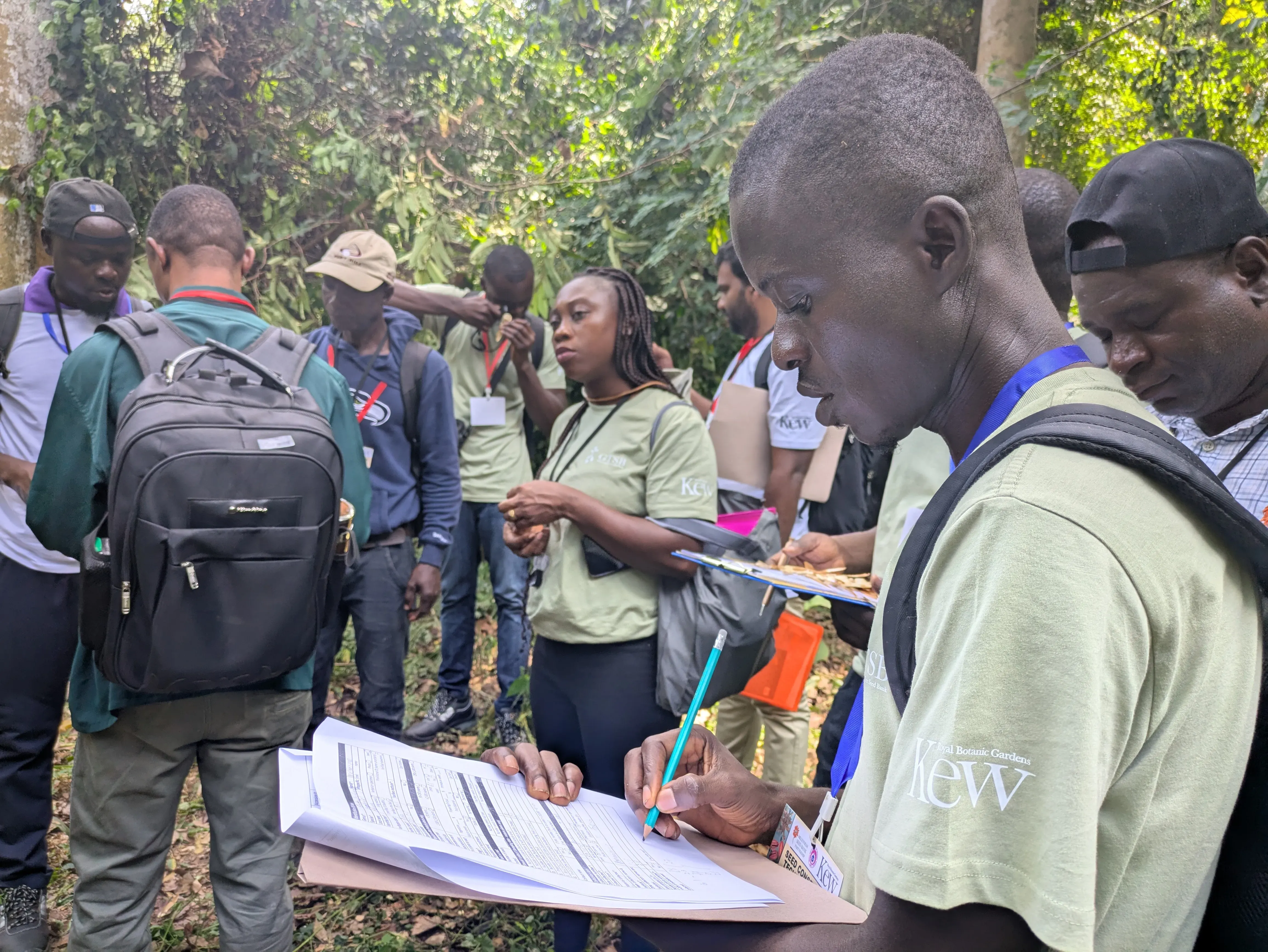 A group of individuals stand in a forest area. They're holding clipboards and discussing seed conservation techniques