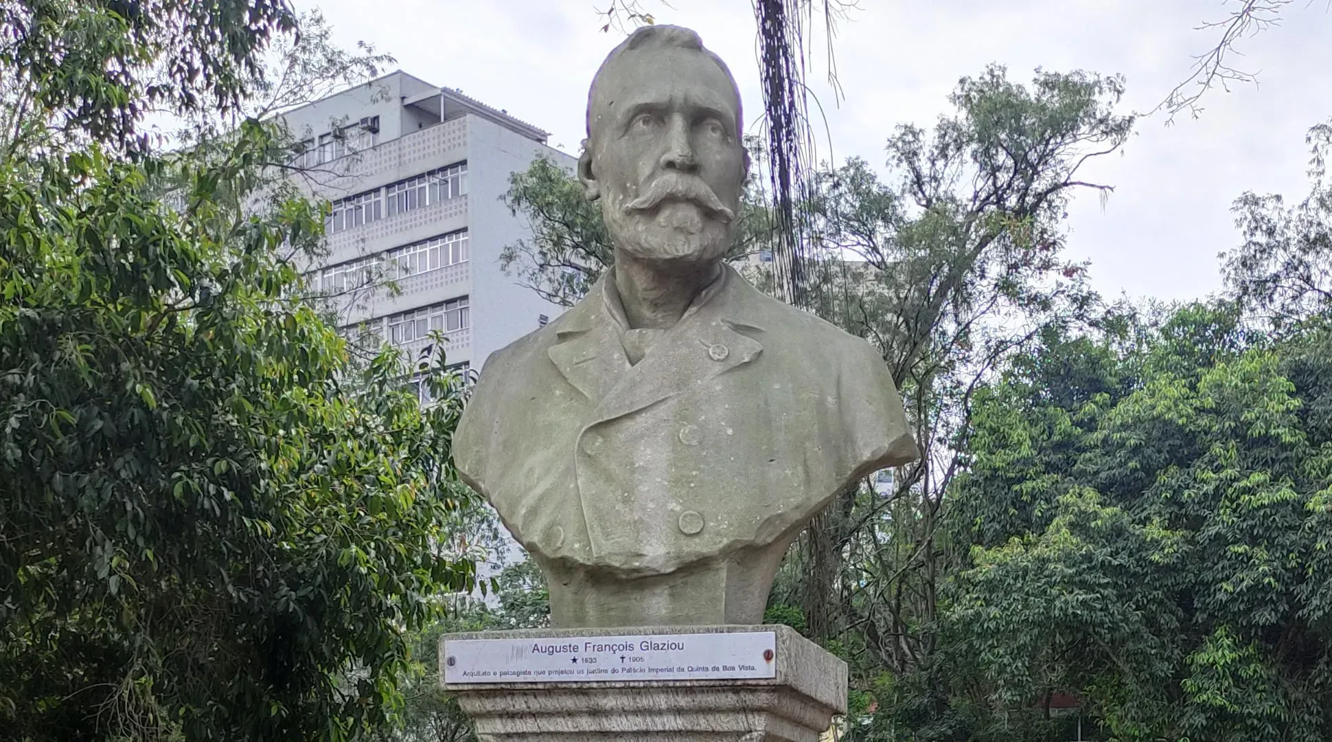A stone bust of a bearded man mounted on a pedestal in a leafy outdoor setting, with trees and a building visible in the background.