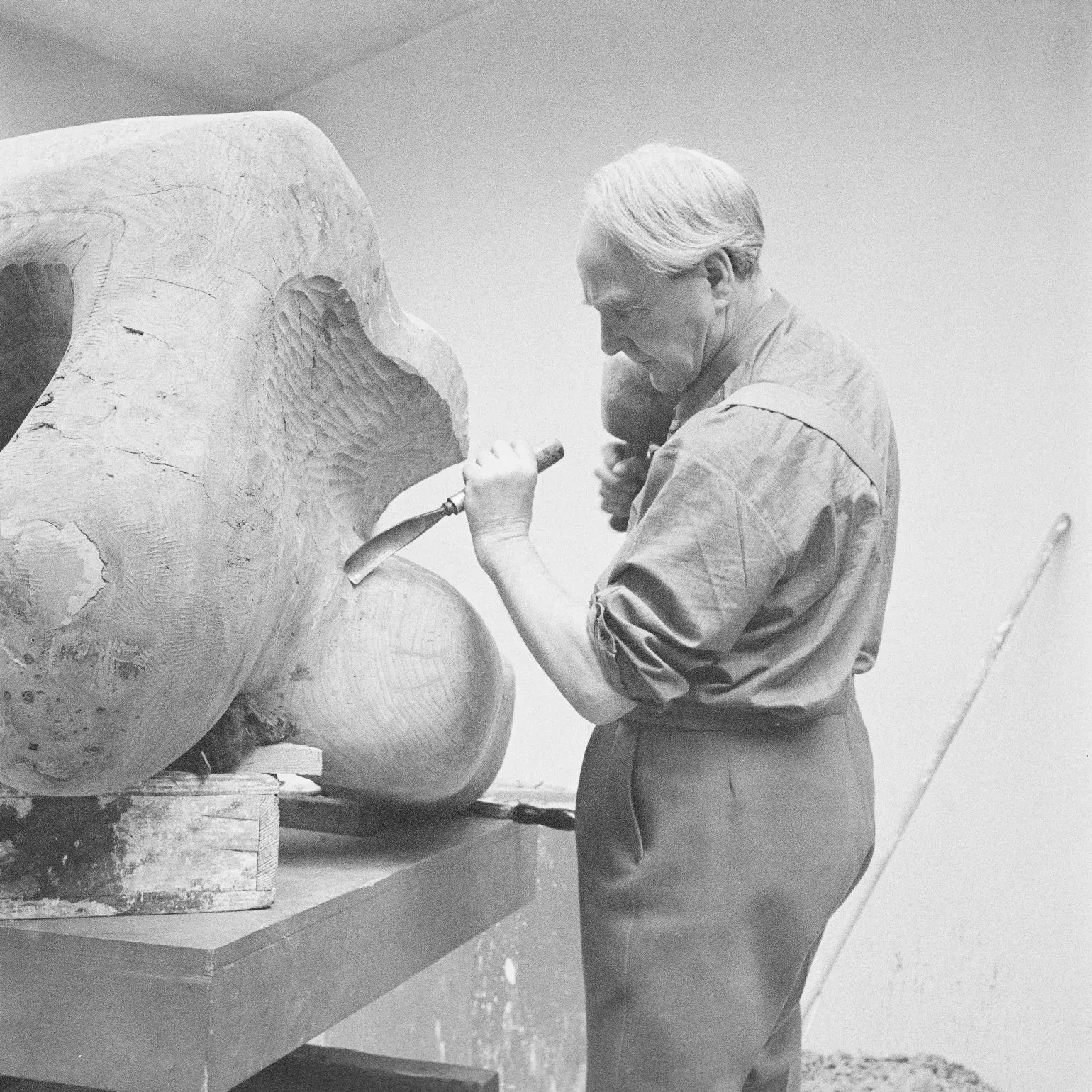 Photograph of Henry Moore standing up carving Reclining Figure in elm wood on a table