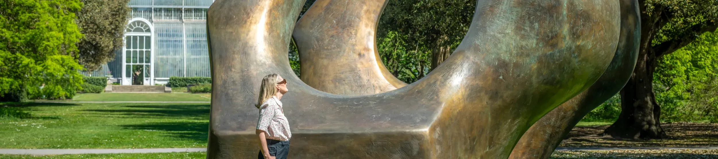 Visitor interacting with Henry Moore's bronze sculpture Double Oval, with a view of the Palm House greenhouse behind it