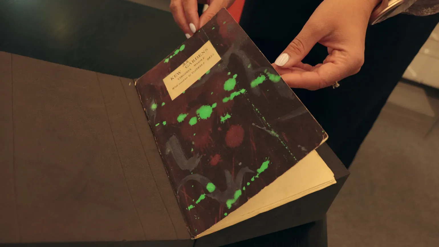 Two hands hold open an old black book cover decorated with paint splodges of green, red and white. Cover reads Kew Gardens, Virginia Woolf, with woodcuts by Vanessa Bell