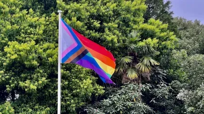 A pride flag, with rainbow colours and a wedge with pink light blue and white flying in front of a variety of green trees