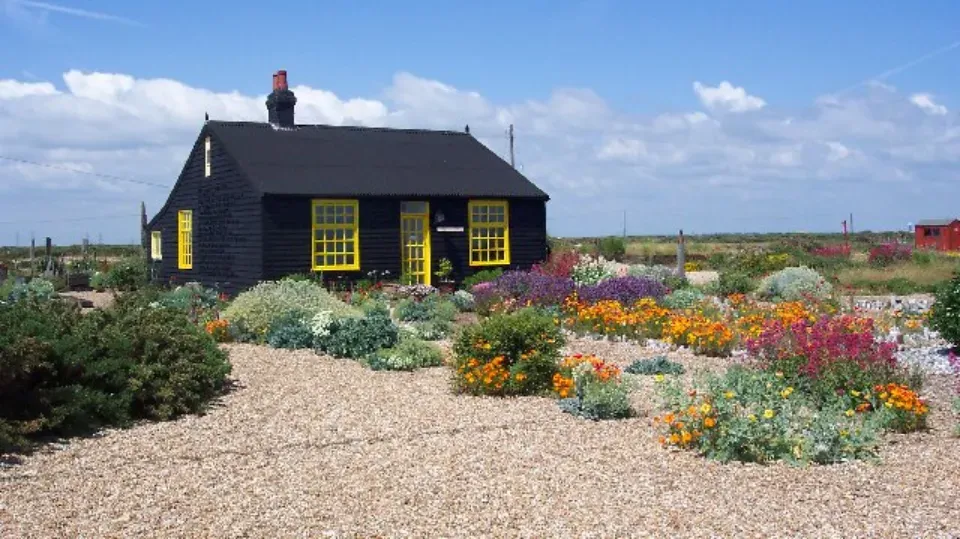 A cottage backed pitch black with a colourful garden growing on shingles under a blue sky
