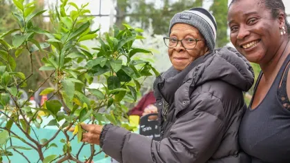 Two Black women, one older and one middle aged, smiling next to a young tree in a conservatory