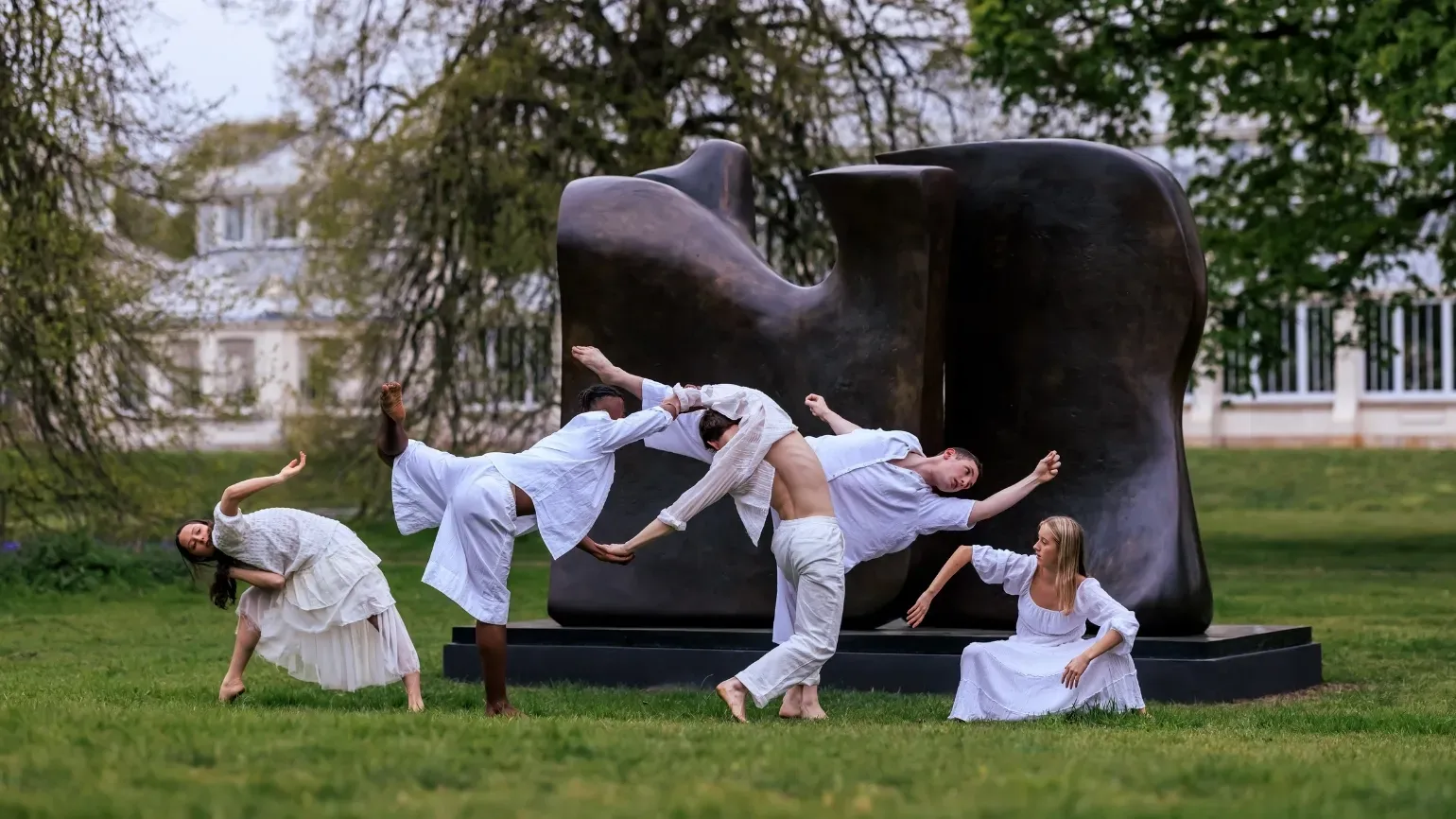 Five people in white clothing, dancing in front of a Henry Moore sculpture at sunset. Temperate House is in the background.