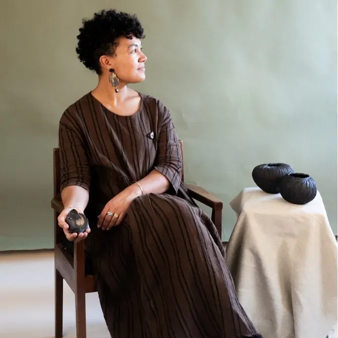 Woman sitting in a chair, her head is turned in profile. She is holding a black-coloured ceramic in her hand.