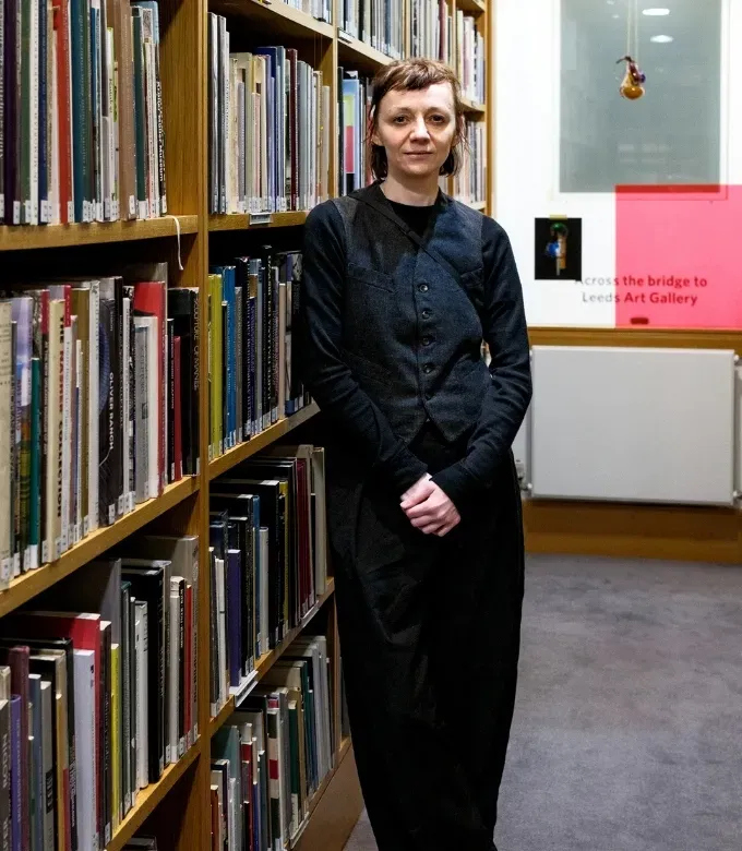Women standing in front of a tall bookcase, in a library aisle. She's dressed in an all black outfit and smiling subtly.