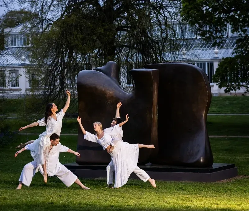 Group of dancers dressed in white, dancing in front of Knife Edge Two Piece by Henry Moore.